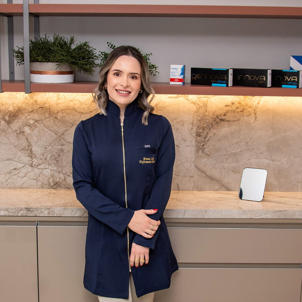 Uma mulher sorridente em um ambiente profissional, usando um jaleco, posando ao lado de uma bancada.