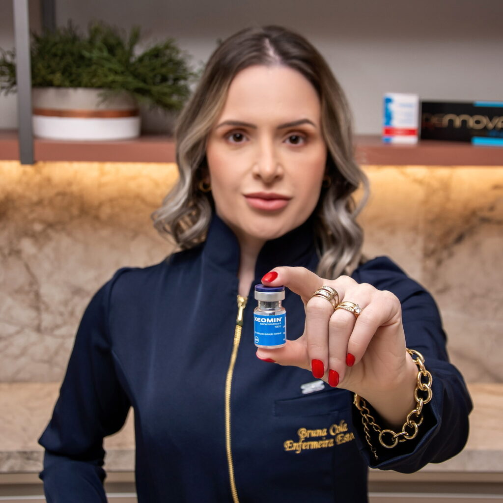 Uma mulher segura um pequeno frasco azul, vestindo um uniforme escuro, em um ambiente moderno.